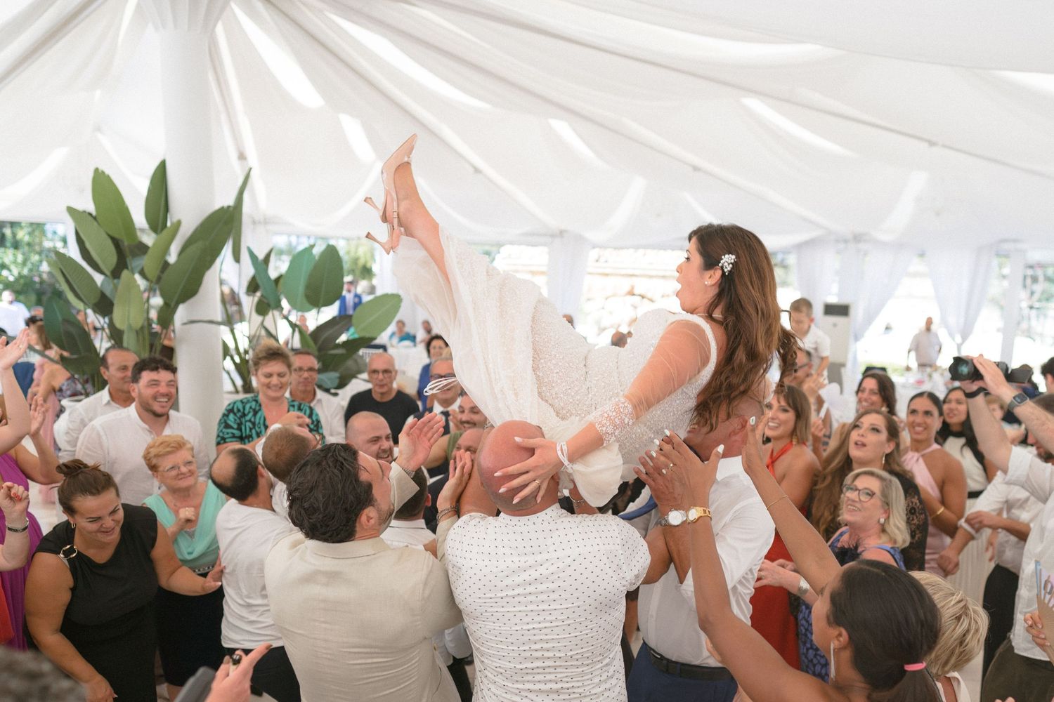 Wedding reception celebration under white tent with guests dancing and cheering.