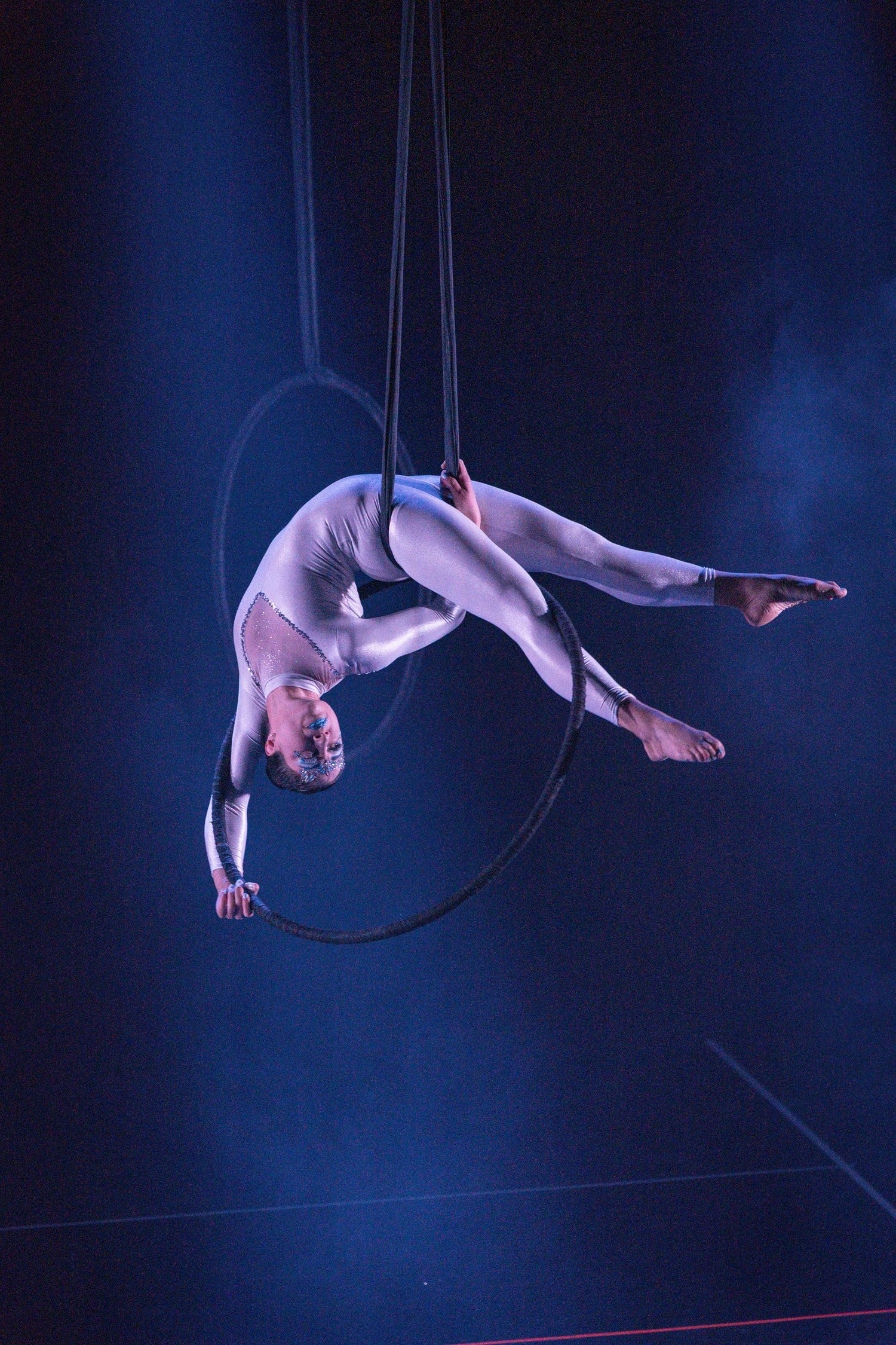 Aerial performer in white costume gracefully suspended on lyra hoop against dark blue dramatic stage lighting.