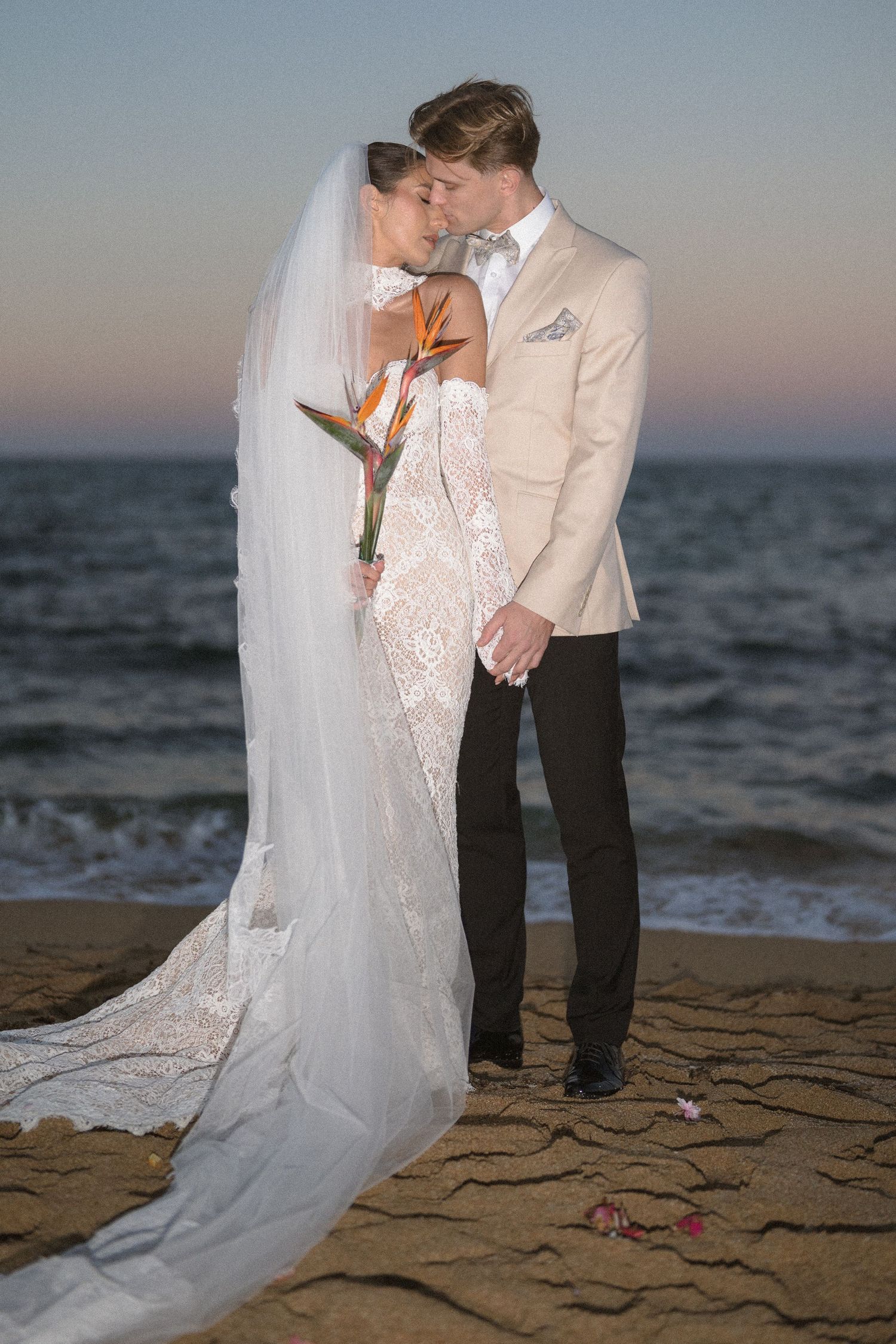 Bride and groom embrace on a beach at sunset, she in a lace gown and long veil holding a bird of paradise bouquet.