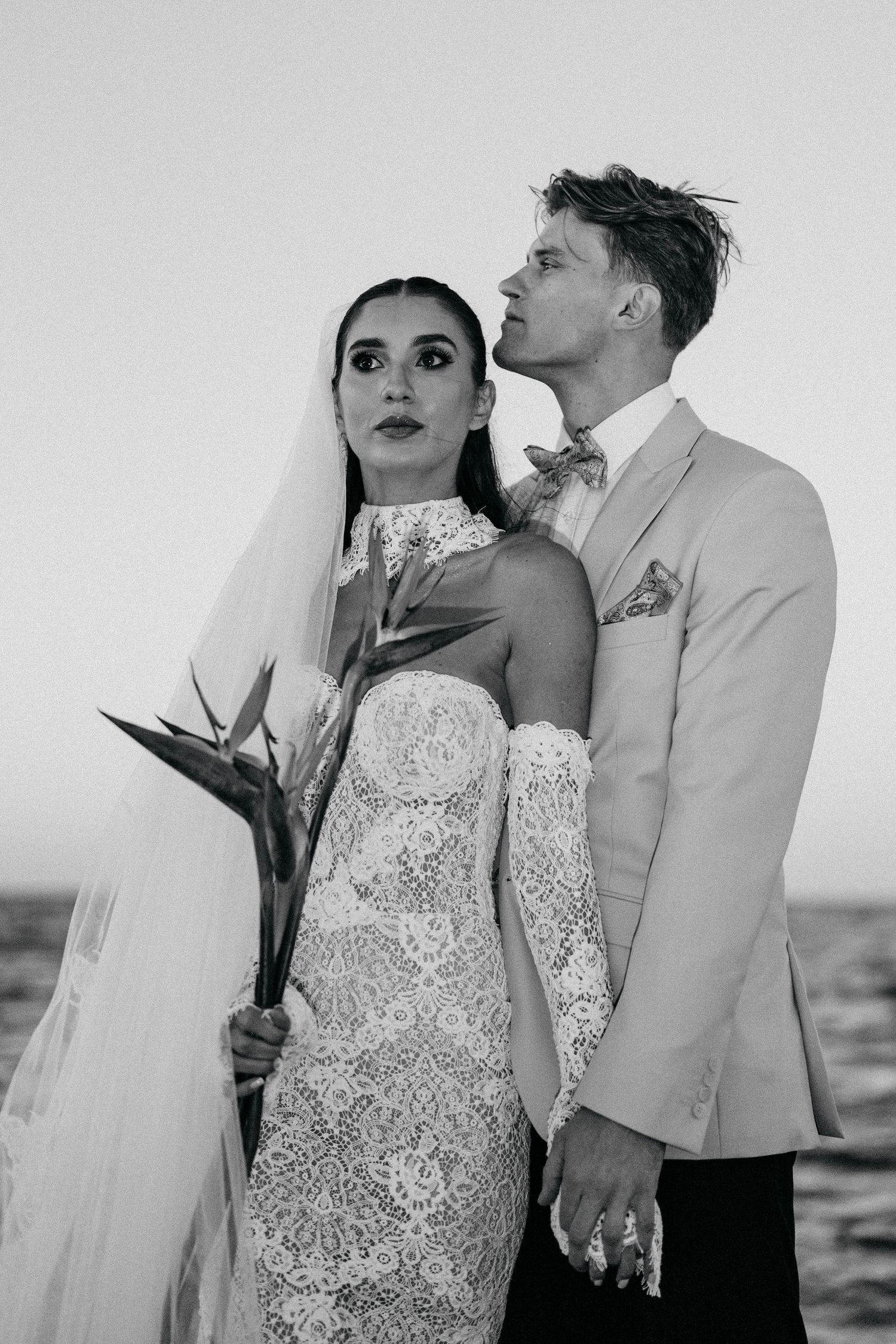 Black and white photo of a bride holding a bird of paradise bouquet beside a groom in a light suit and bow tie.