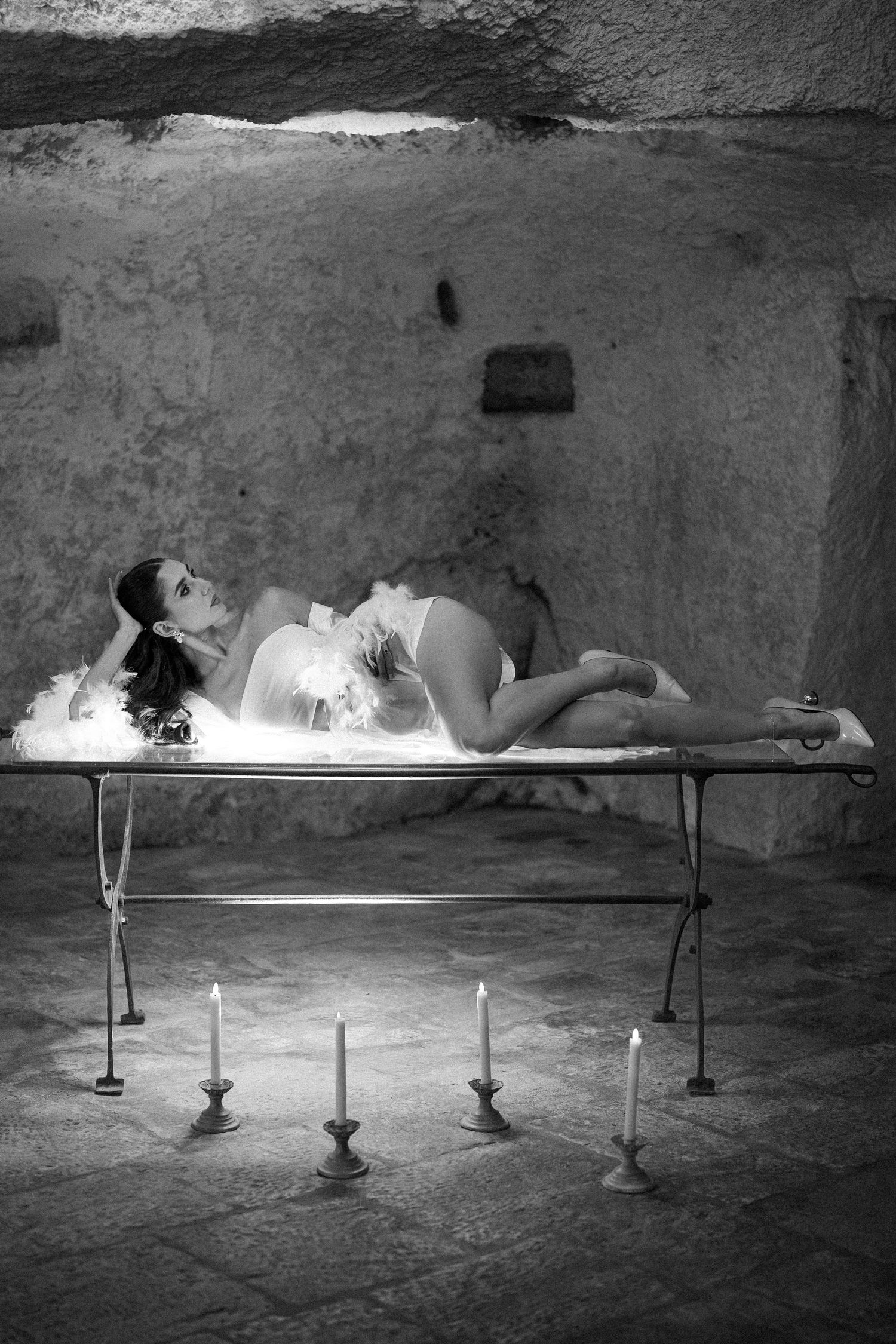 Black and white image of a bride lying on a table with feathered white skirt in a candlelit stone room.