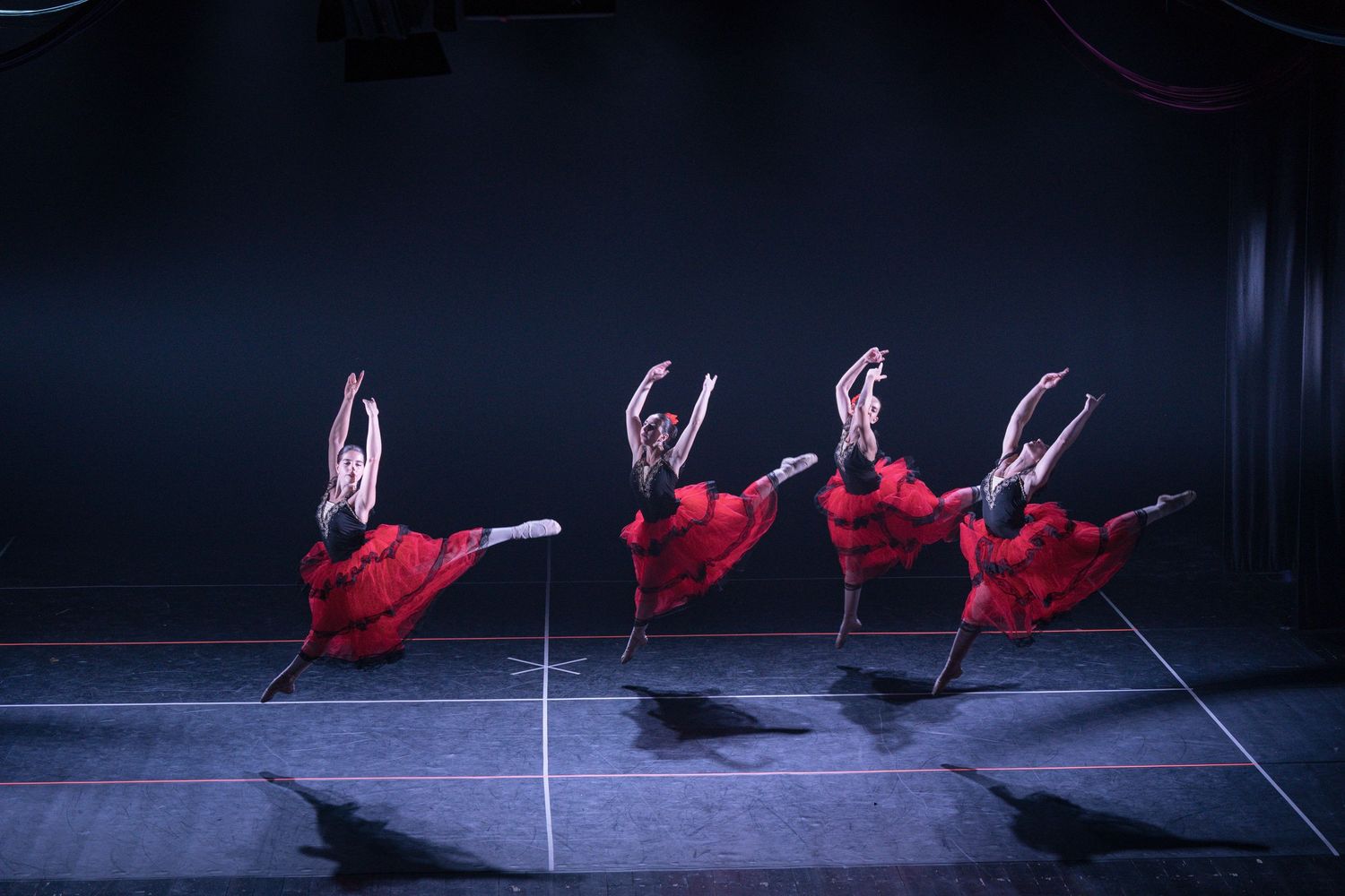 Ballet dancers in flowing red dresses perform synchronized leaps across a dark stage during a classical performance.