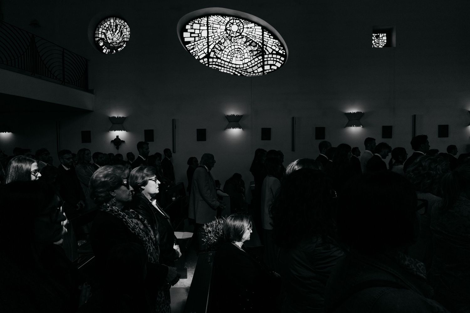 Wedding guests gathered in dimly lit church with ornate stained glass ceiling in black and white.