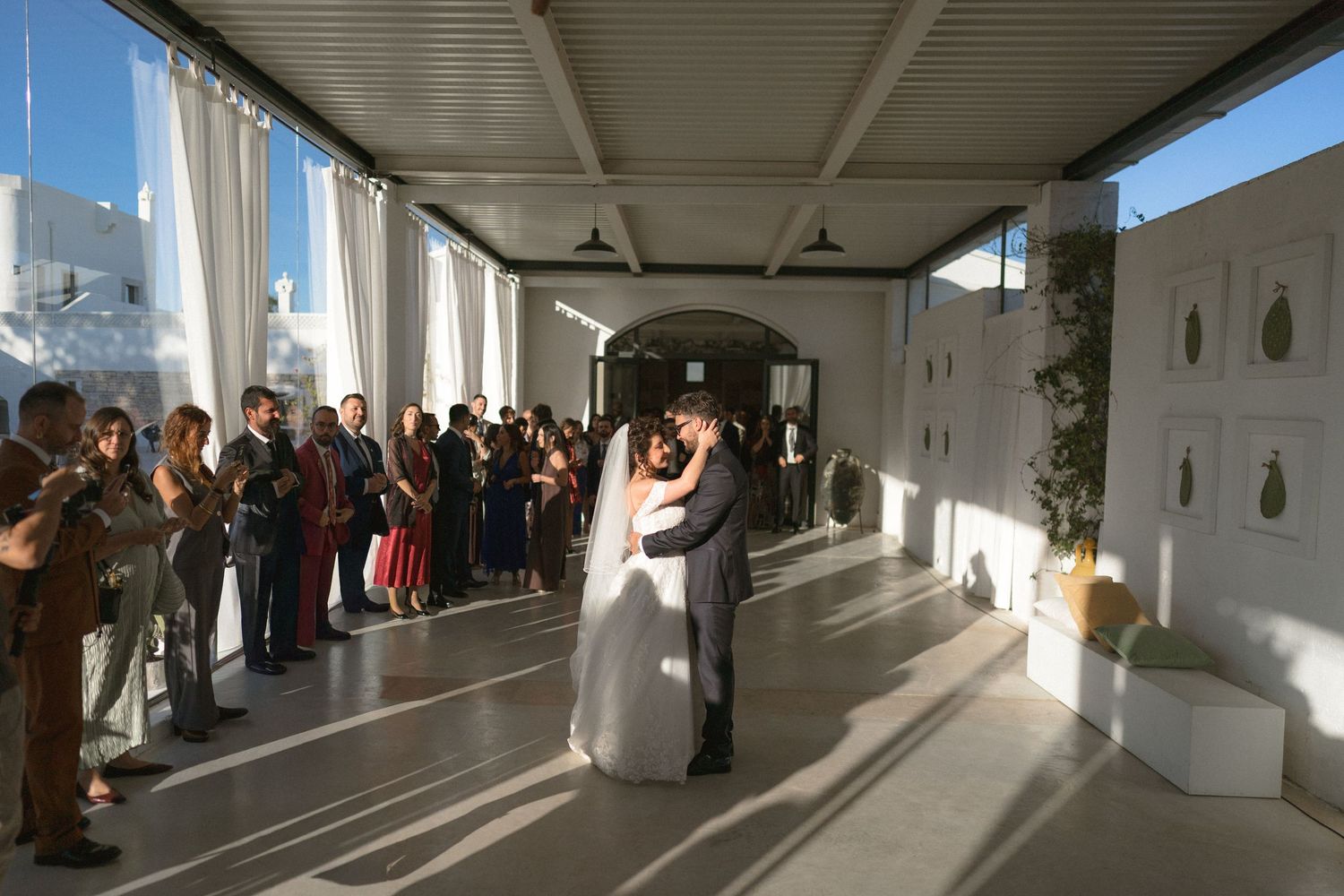 Bride and groom share first dance in bright airy venue while wedding guests applaud and celebrate.