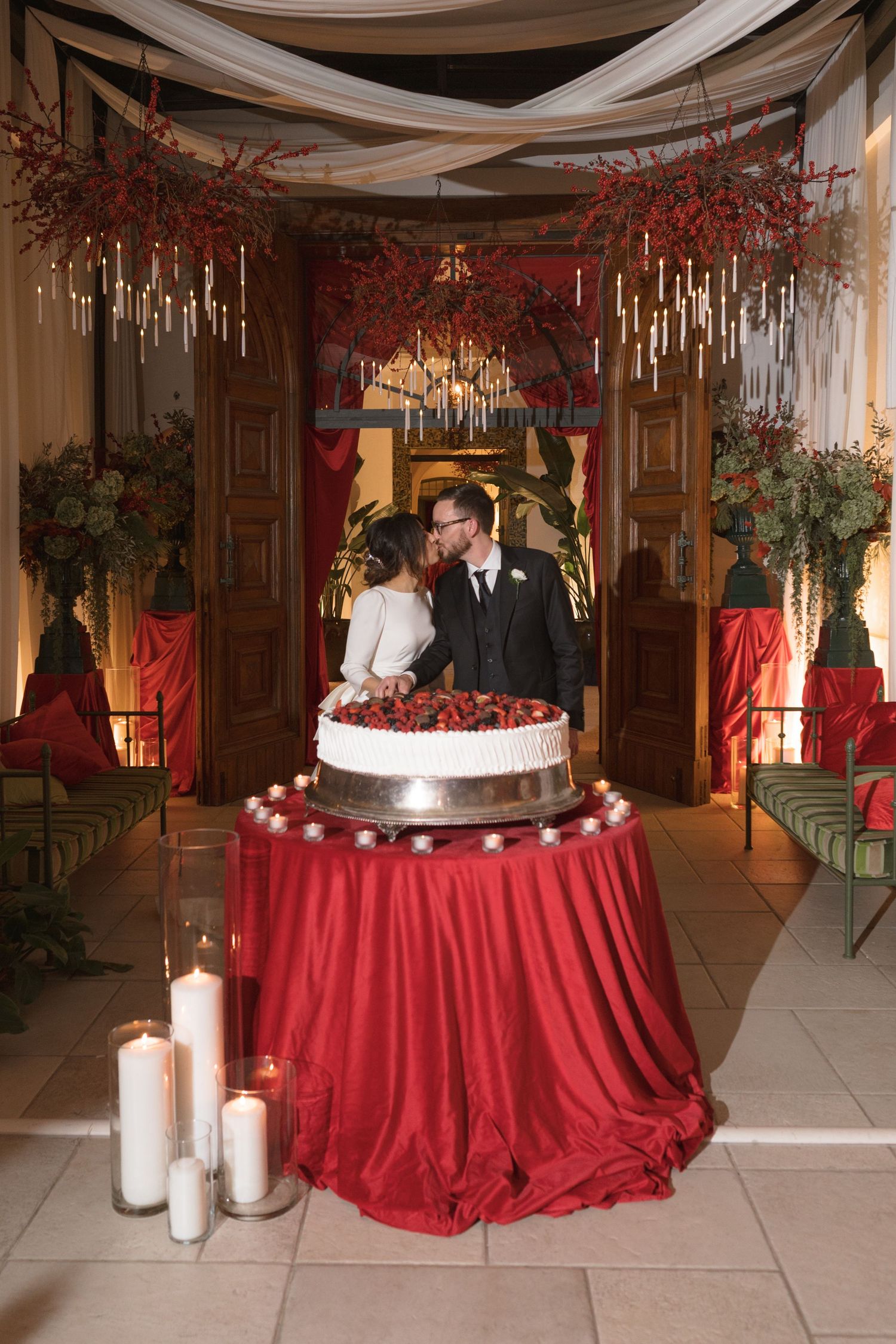 Newlyweds kissing behind white wedding cake on red-draped table surrounded by candles and floral decor.
