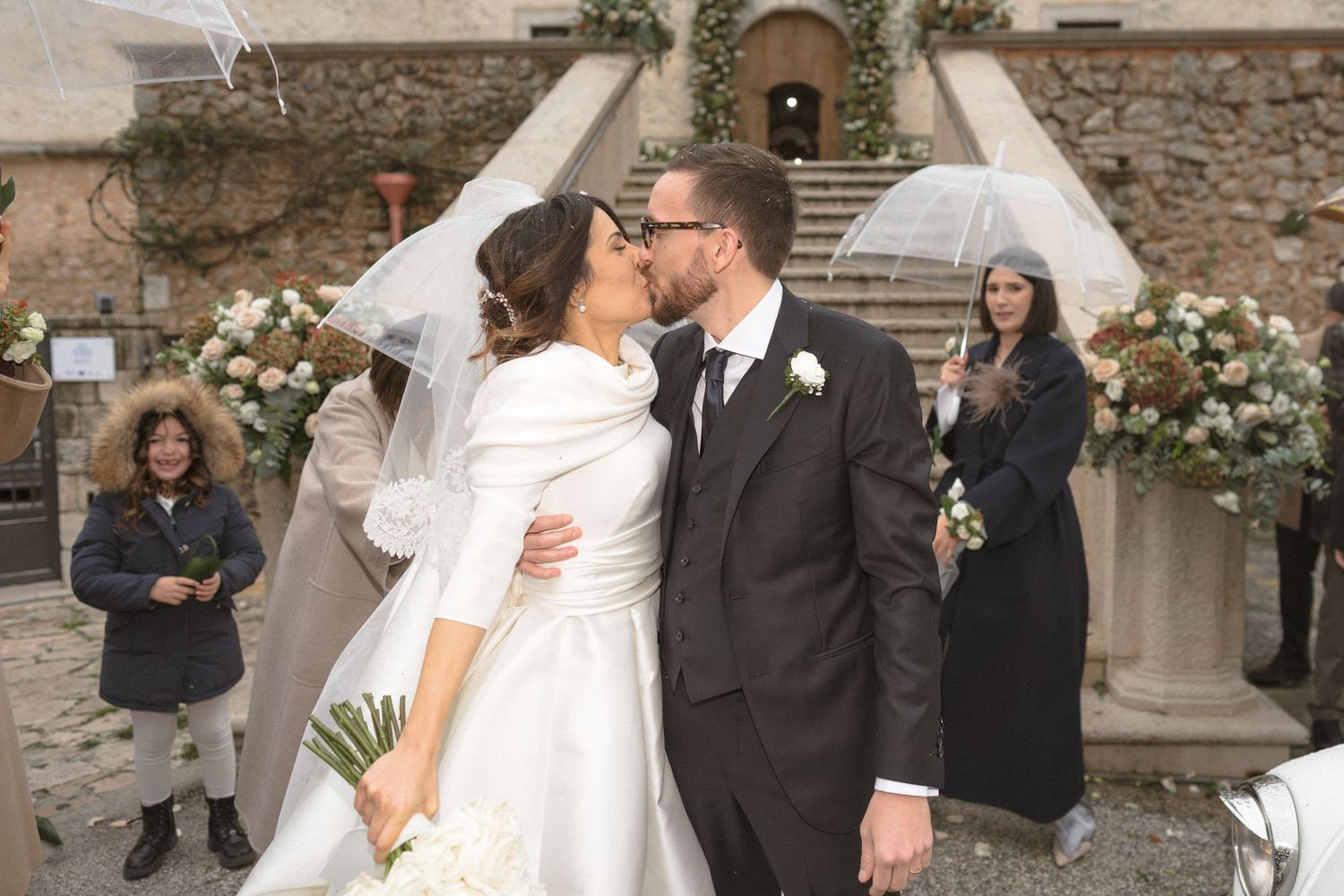 Bride and groom share first kiss outside stone venue with floral decorations on winter wedding day.
