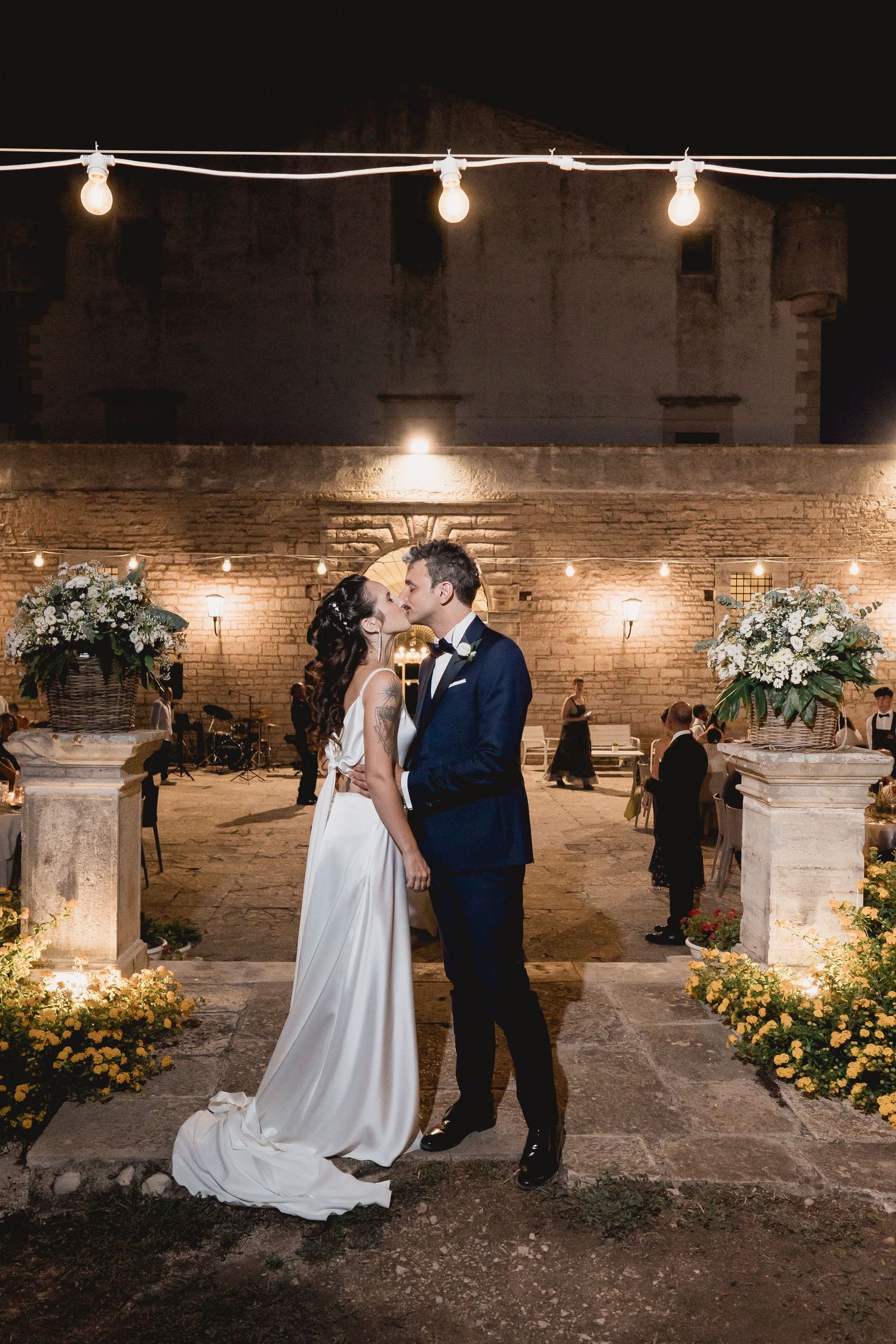 A romantic wedding couple shares a kiss under string lights and floral decor against a rustic brick wall.