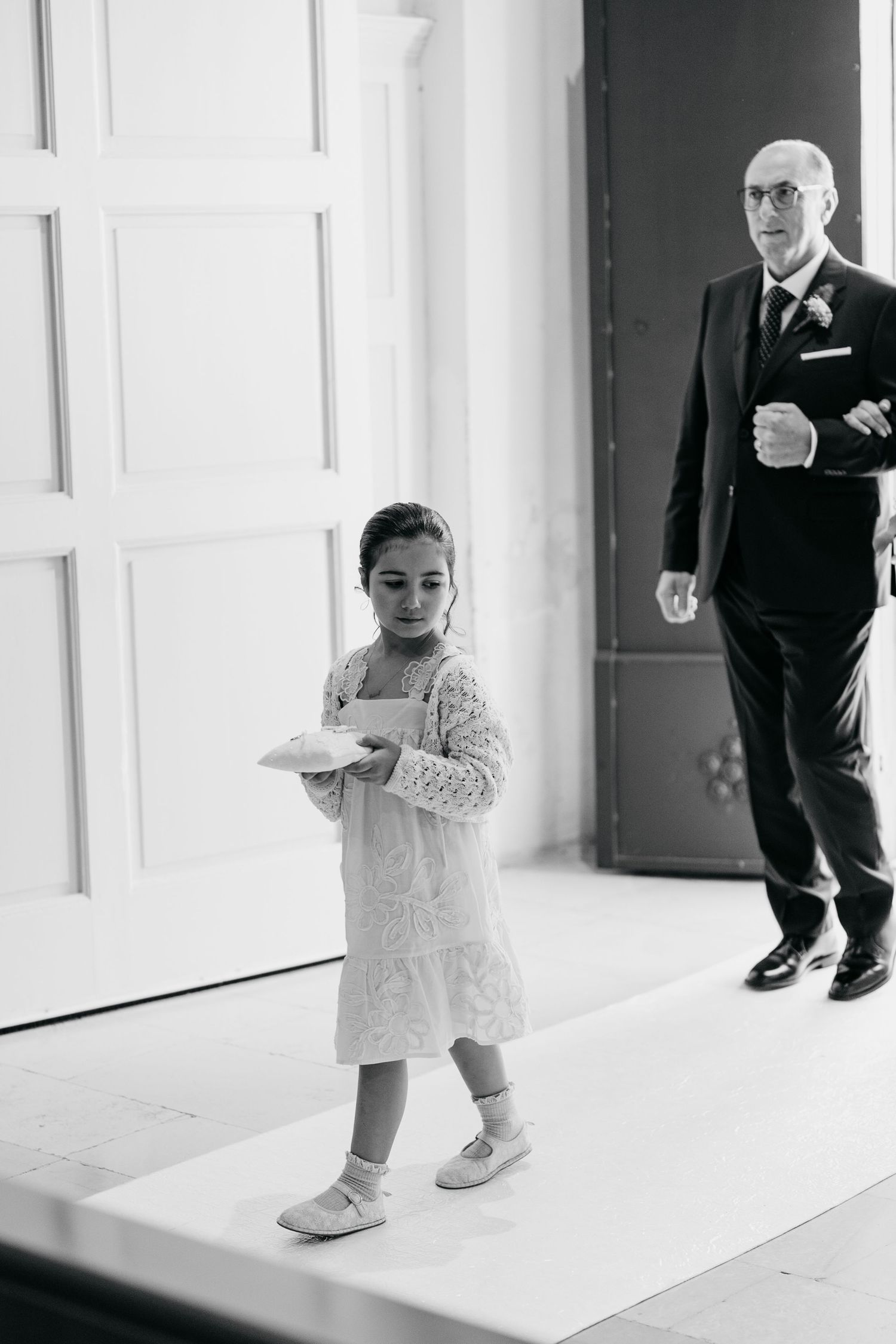 Black and white photo of a small child in formal wear walking down a hallway.