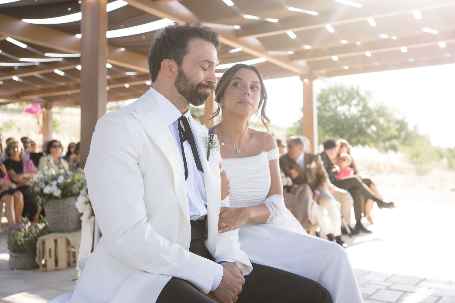 Couple seated on wooden stage during outdoor wedding ceremony with guests visible in background.
