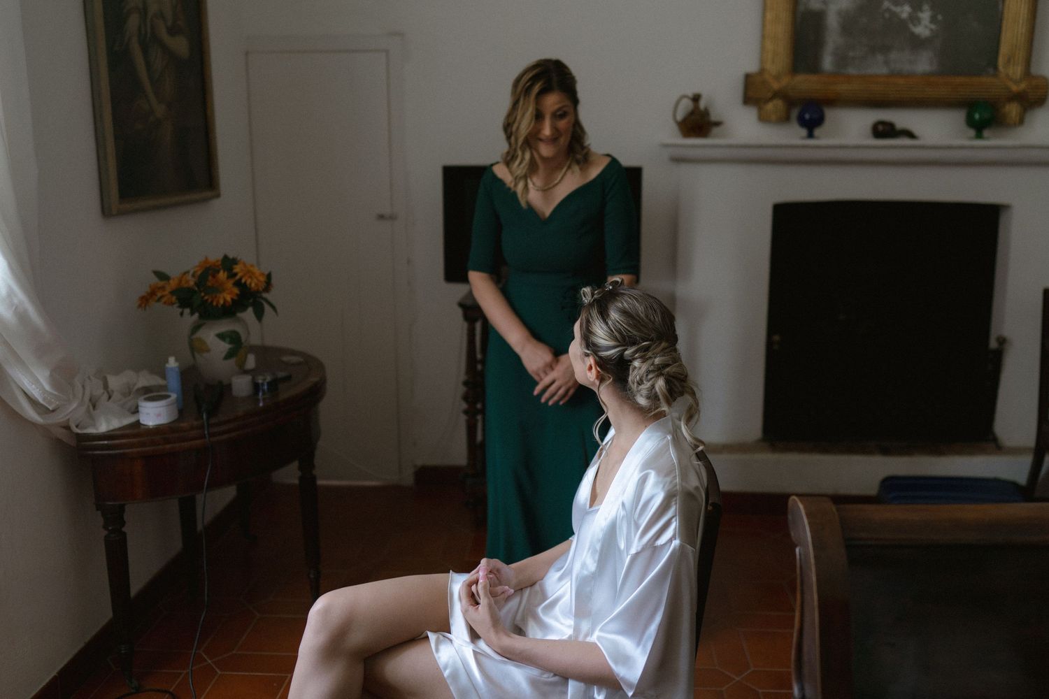 Woman in white robe sitting on floor while getting hair styled in sunlit room.