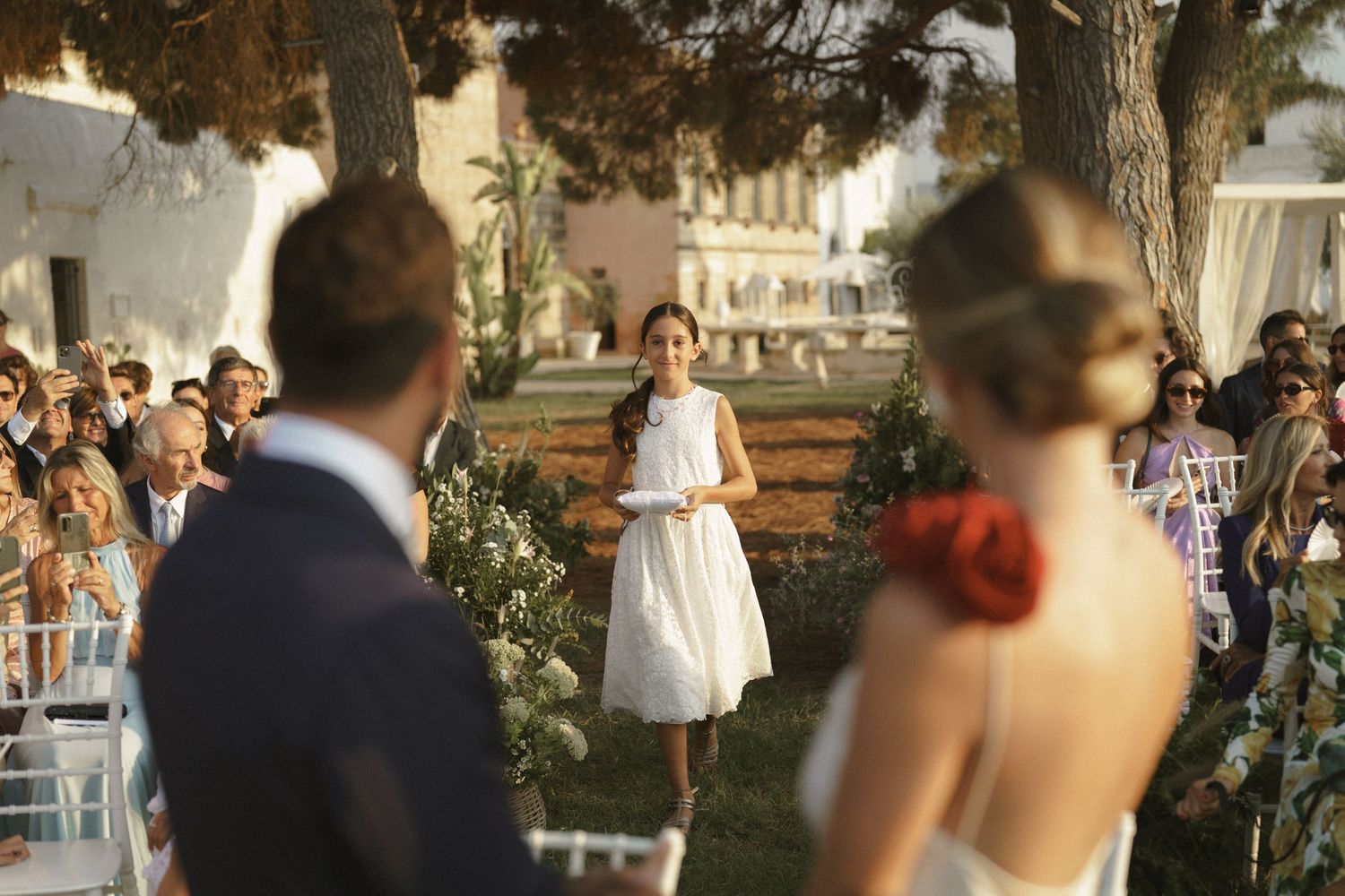 An outdoor wedding ceremony in a garden setting with guests seated on white chairs.