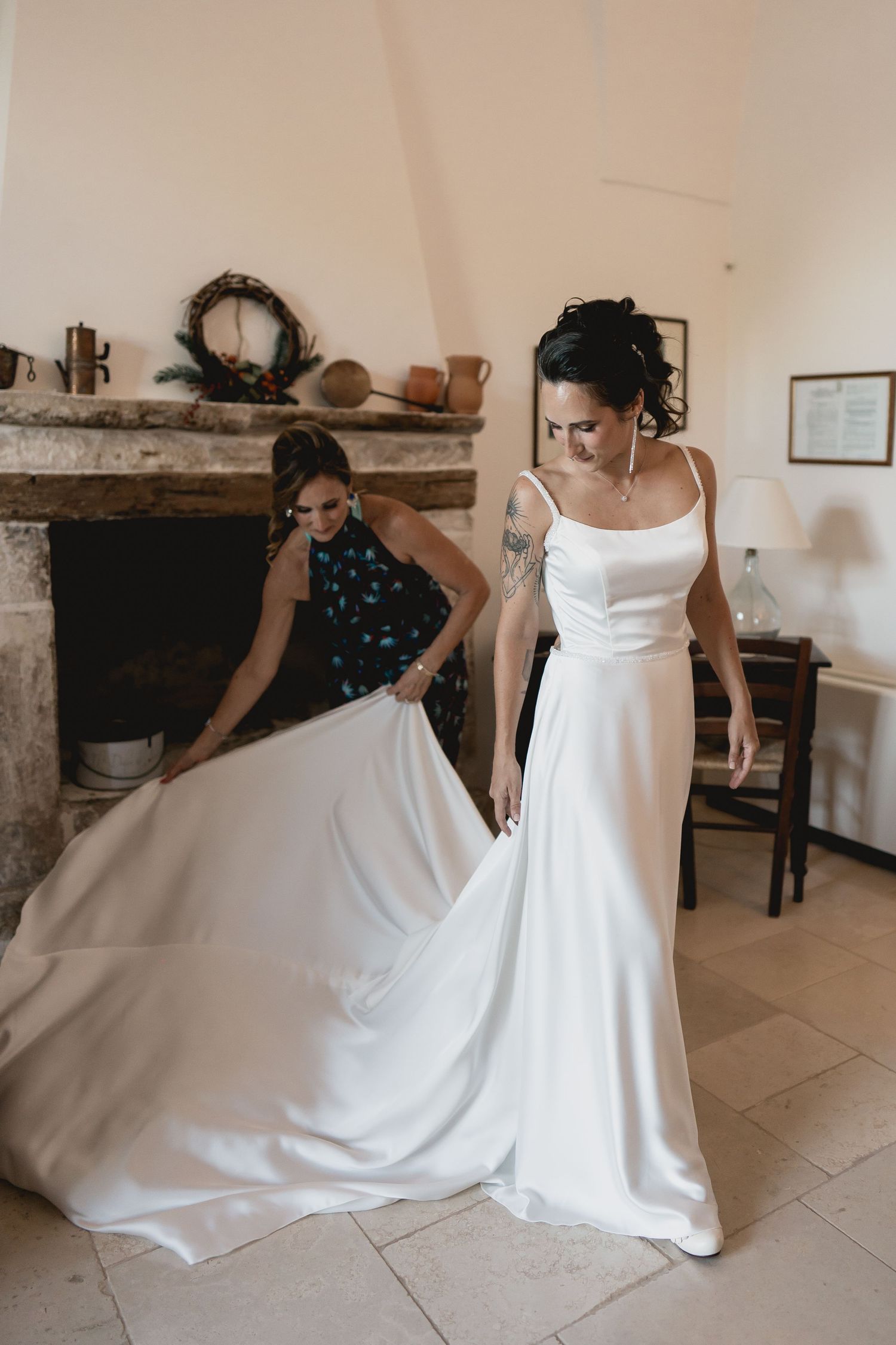 A bride in an elegant white spaghetti strap wedding gown being fitted in a rustic room.