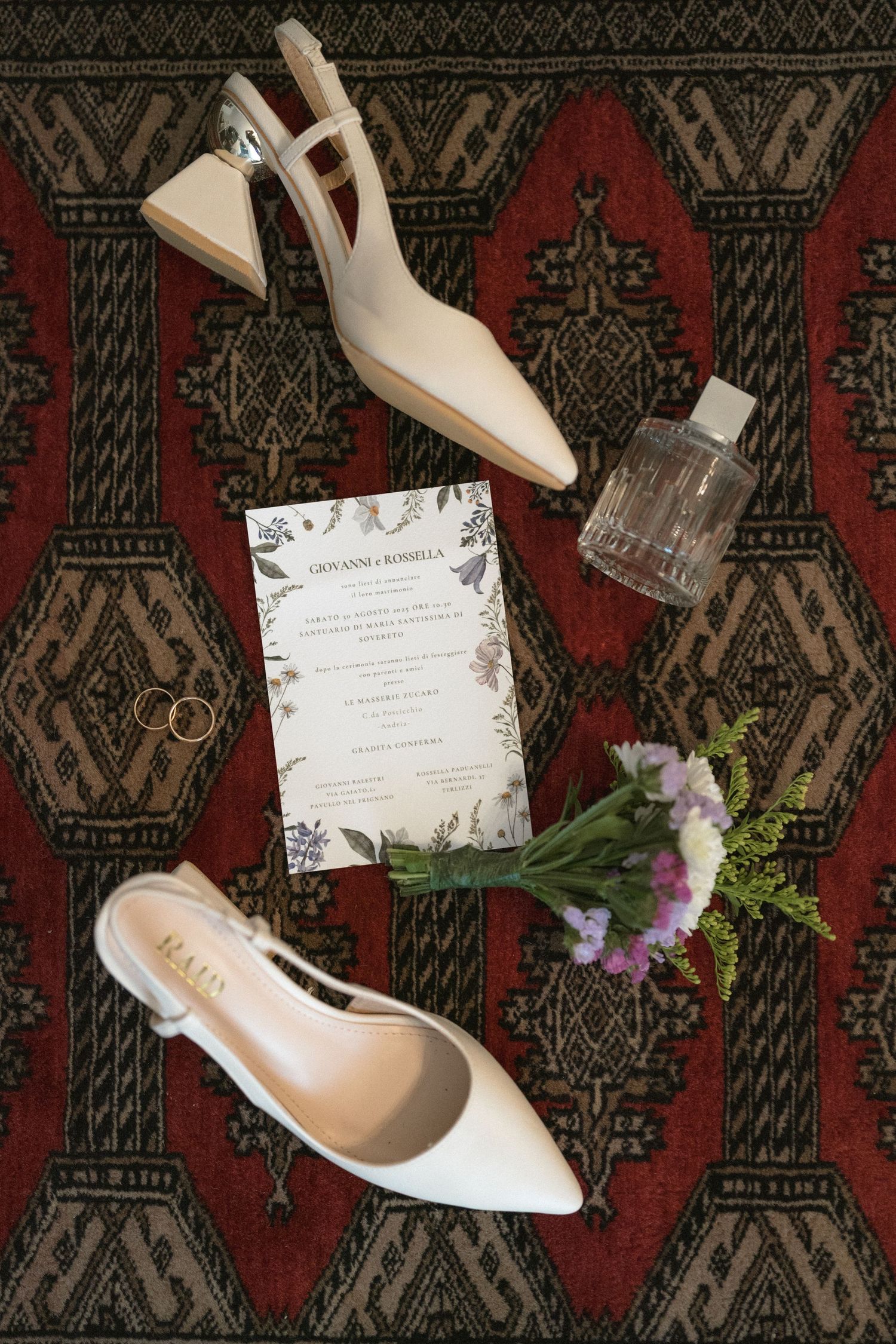 Wedding details flatlay with white shoes, invitation cards and flowers on a patterned rug.