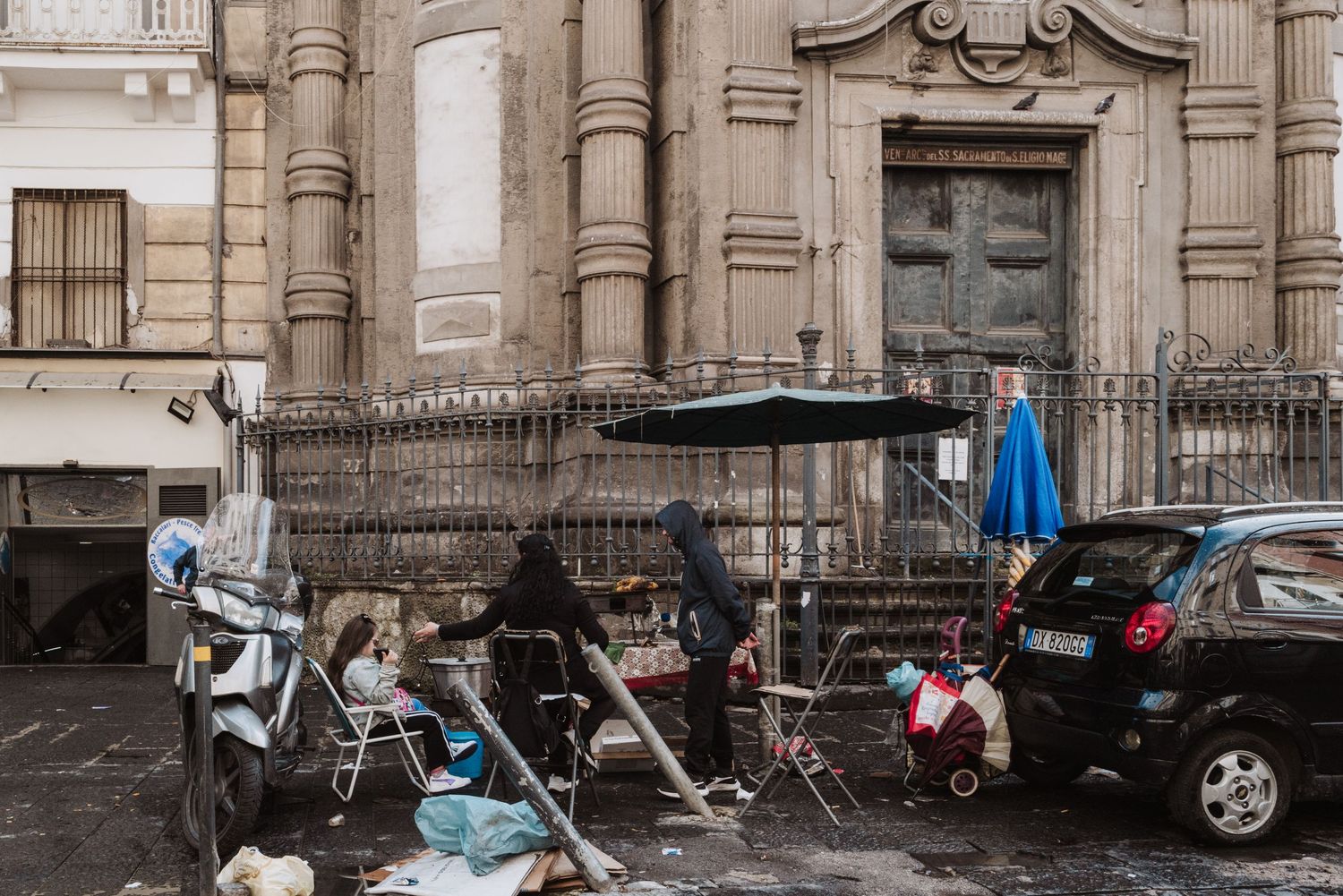 People gathered outside a historic building with cafe chairs and umbrellas on a rainy day in Italy.