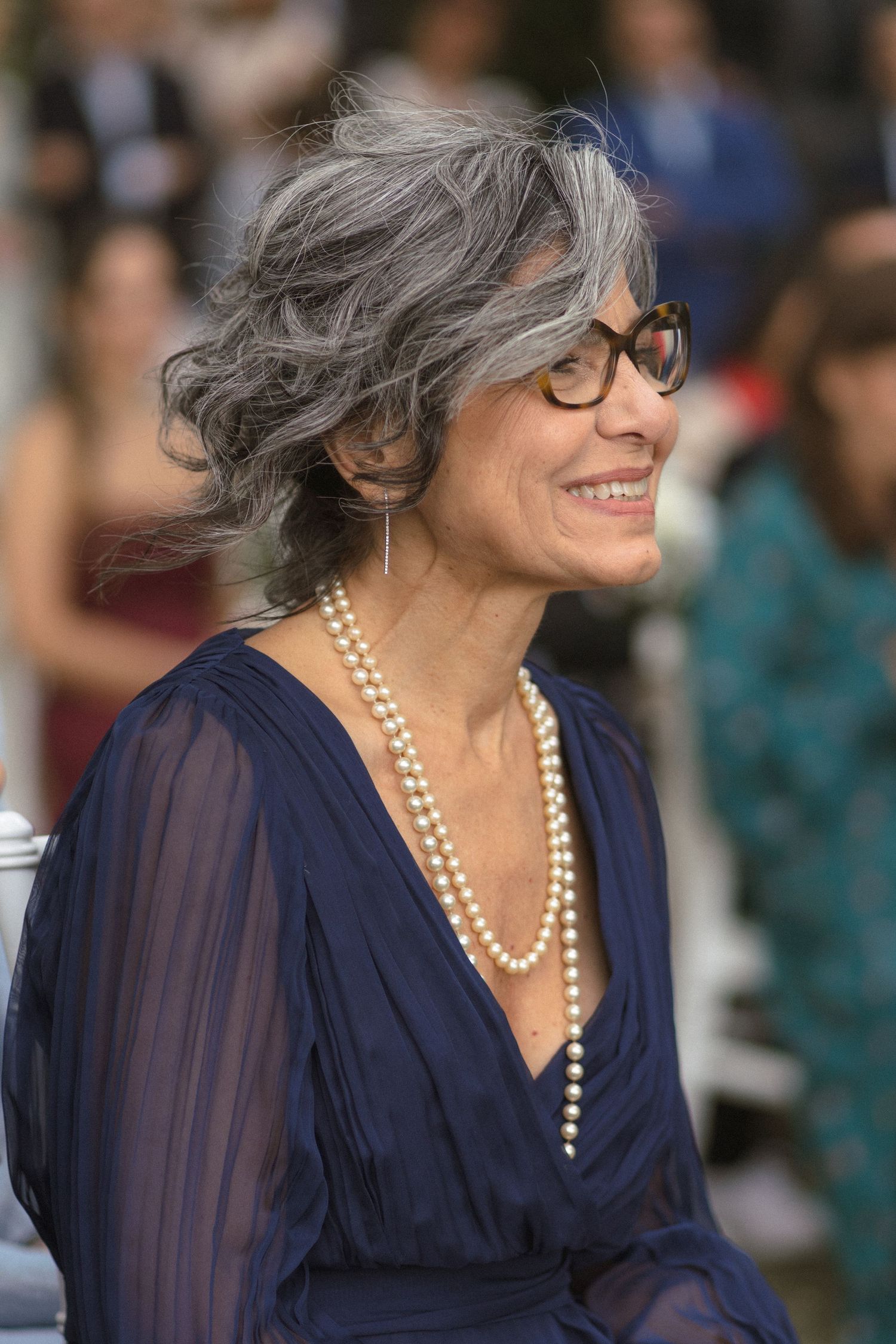 Elegant woman in navy blue dress and pearl necklace smiling at outdoor event.