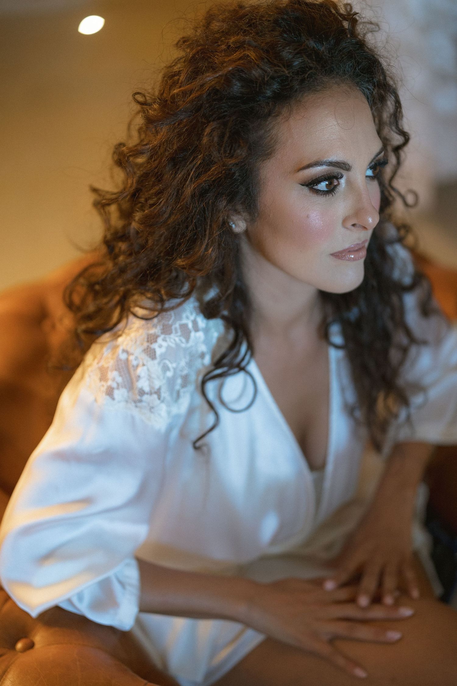 A bride in a white silk robe with curly hair and elegant makeup during wedding preparations.