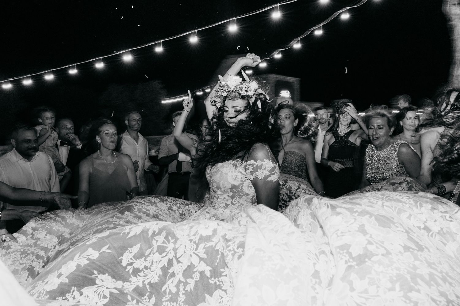 Wedding guests dancing and celebrating on a dance floor under string lights at an evening reception.