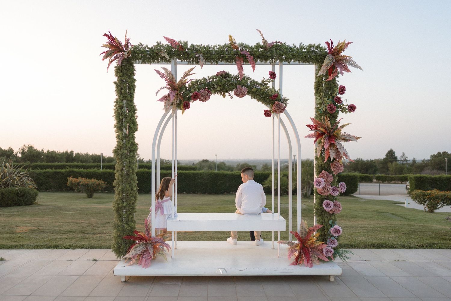 Outdoor wedding ceremony setup with white arch decorated with pink flowers and greenery against a lakeside backdrop.