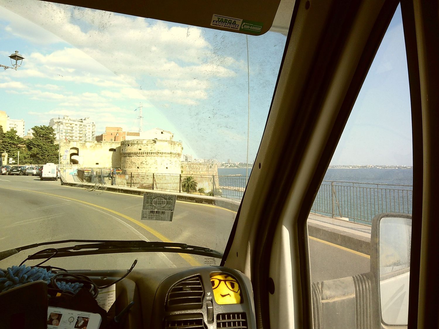 Coastal road view through car windshield with buildings and ocean visible.