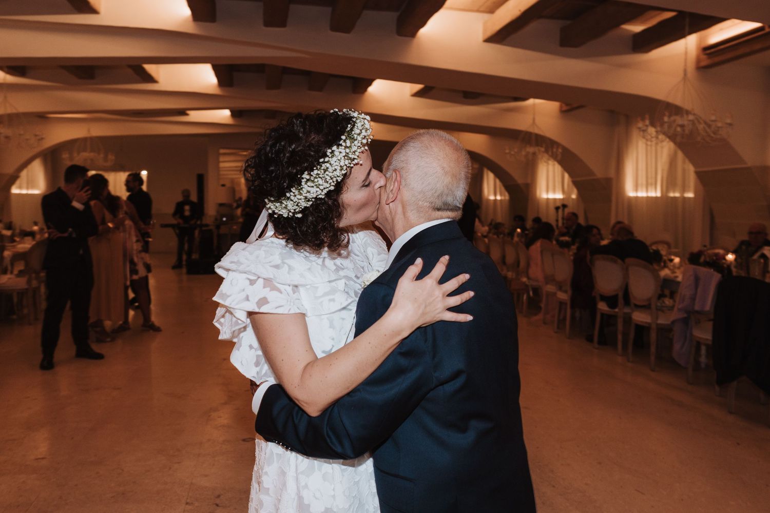 Newlyweds share their first dance in a beautifully lit reception hall with exposed wooden beams.