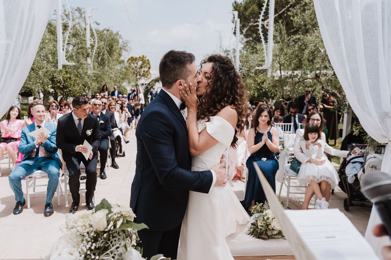 A joyful wedding ceremony moment with guests watching under white drapes and greenery outdoors.
