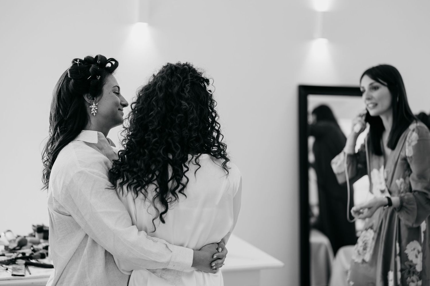 Two people embracing in a bright room with others in the background.