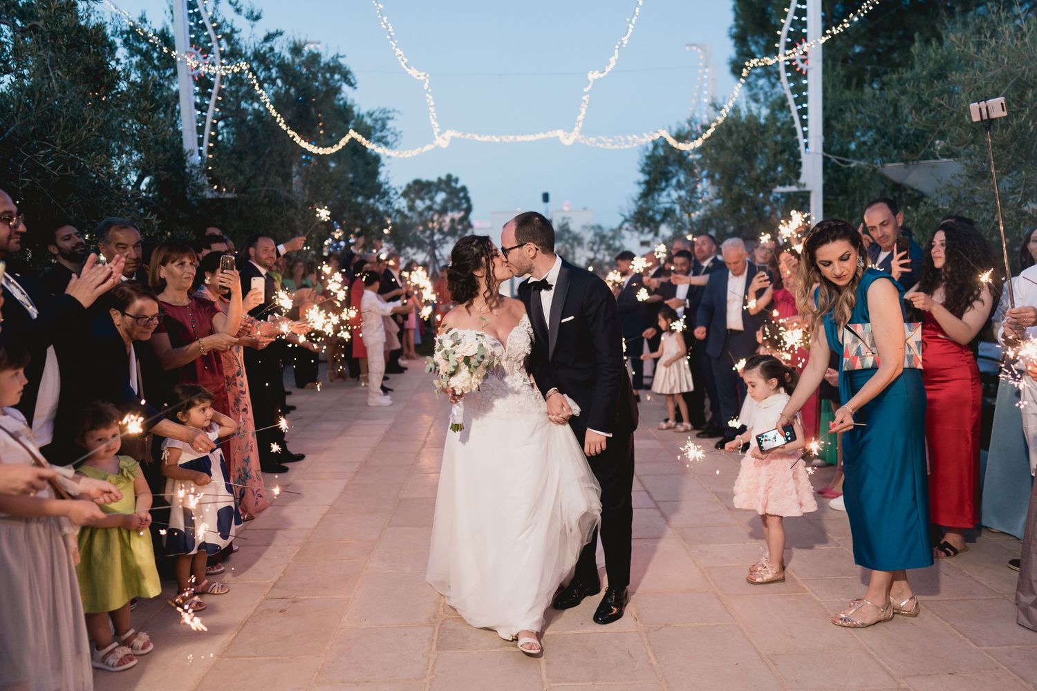 Wedding sparkler exit with guests lining pathway under string lights at dusk.
