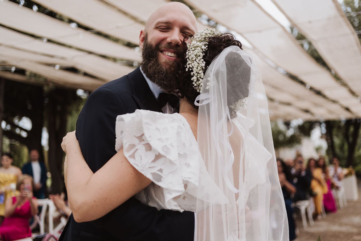 Tender embrace during wedding ceremony under rustic white canopy with guests in background.
