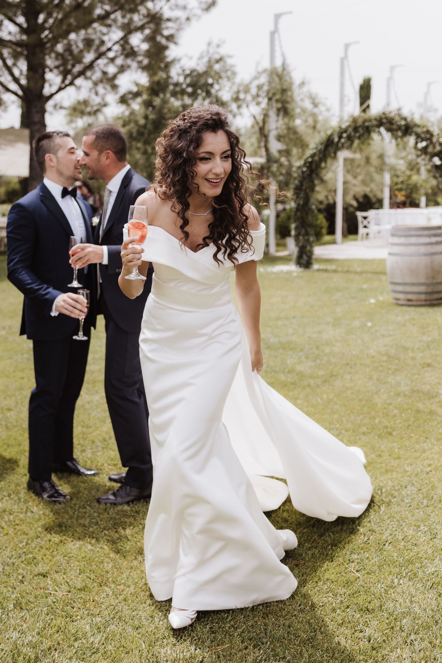 A bride in a fitted white dress walks across a lawn holding a champagne glass.