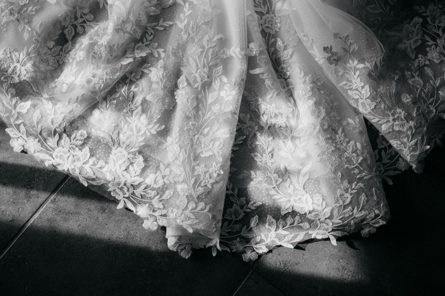 Black and white photo of an elegant lace wedding dress with floral embroidery spread on a tiled floor.