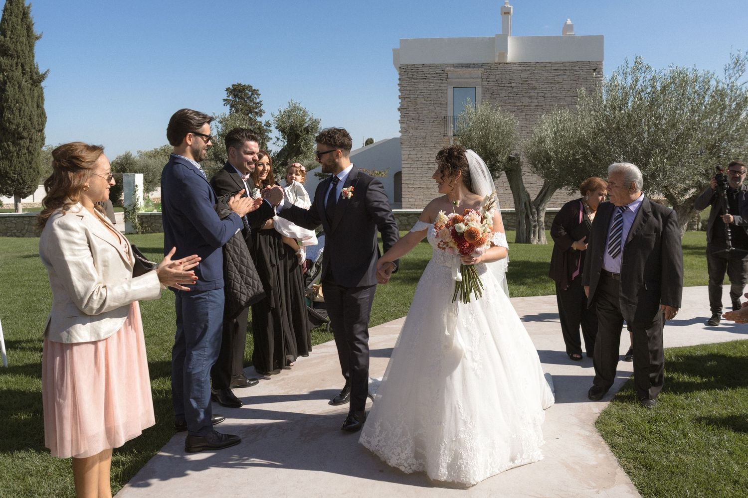 Newlyweds walk through applauding guests on garden path after outdoor wedding ceremony.