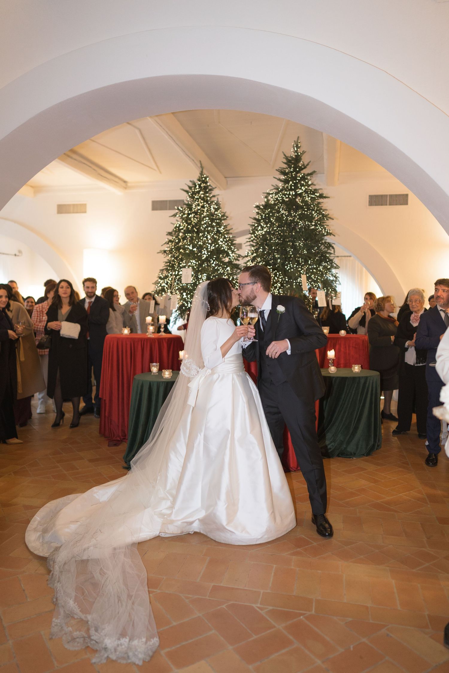 Newlyweds share first kiss on dance floor with Christmas trees and guests in background.