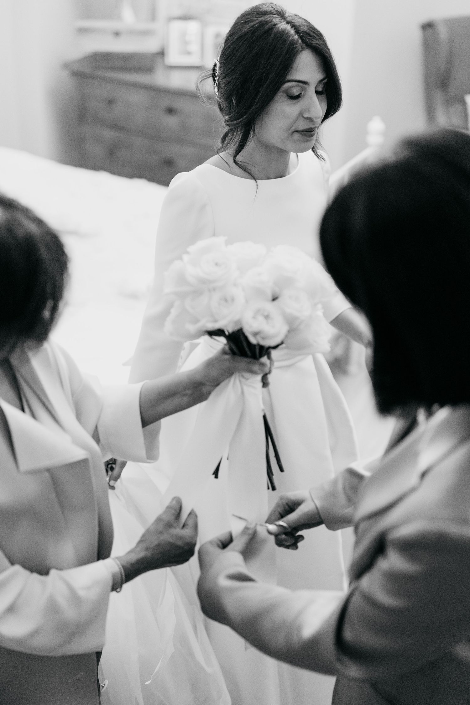 Black and white photo of bride in white gown holding rose bouquet while attendants adjust her dress.