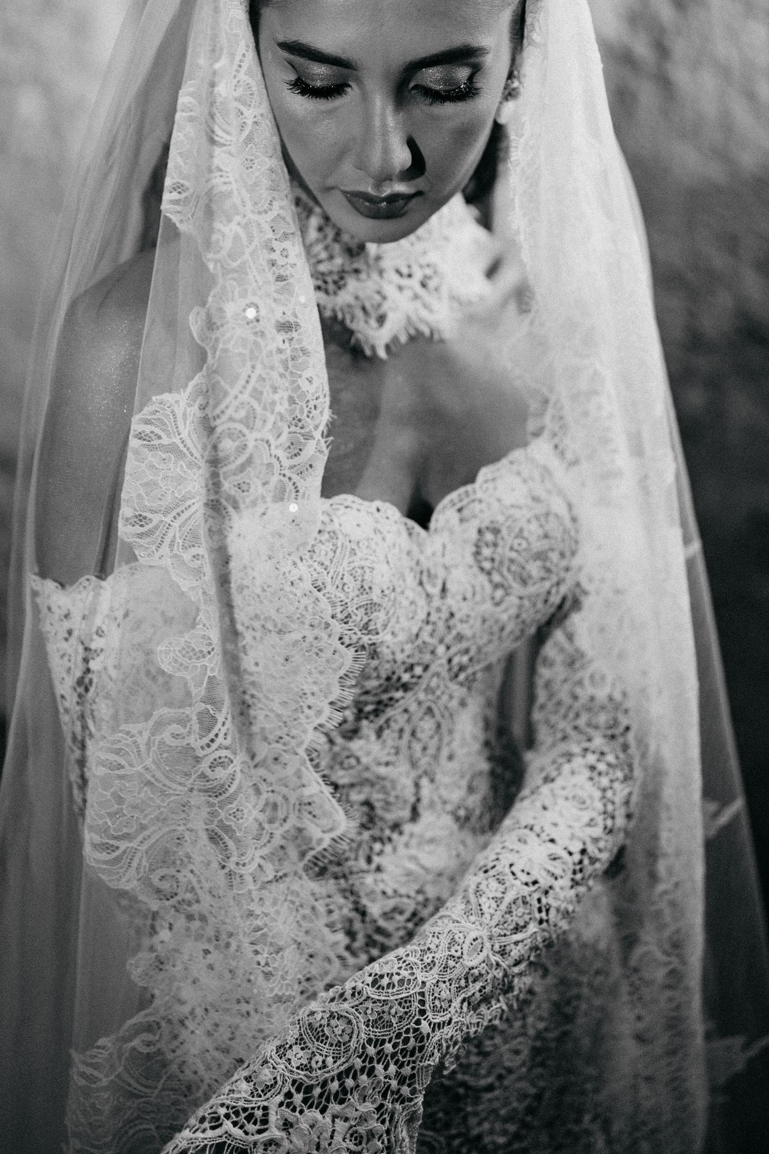 Black and white portrait of a bride looking down, wearing a lace wedding gown and a lace-trimmed veil.