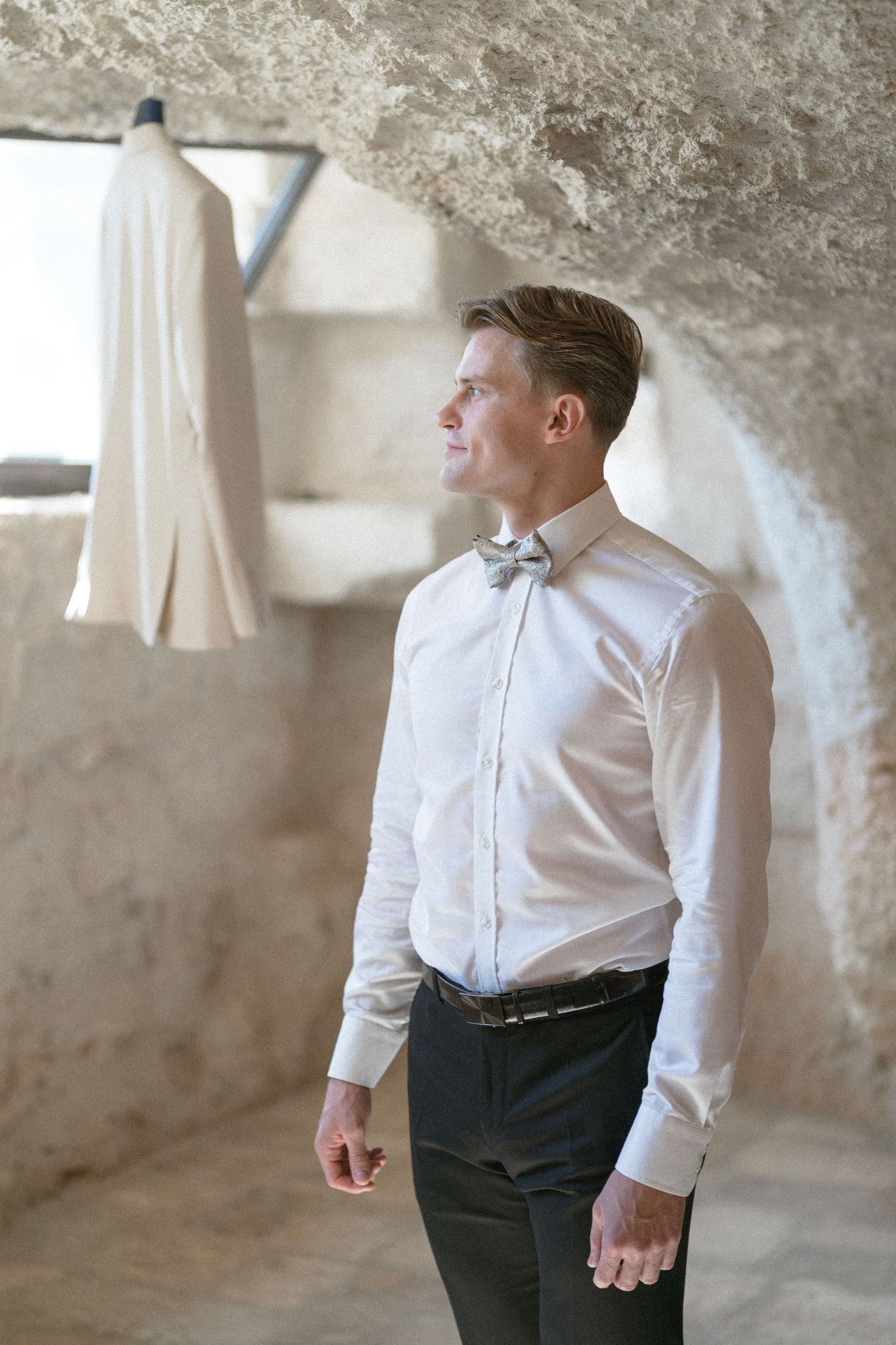 Groom in a white dress shirt and gray bow tie gazes aside inside a rustic stone room, with a cream suit jacket hanging behind him.
