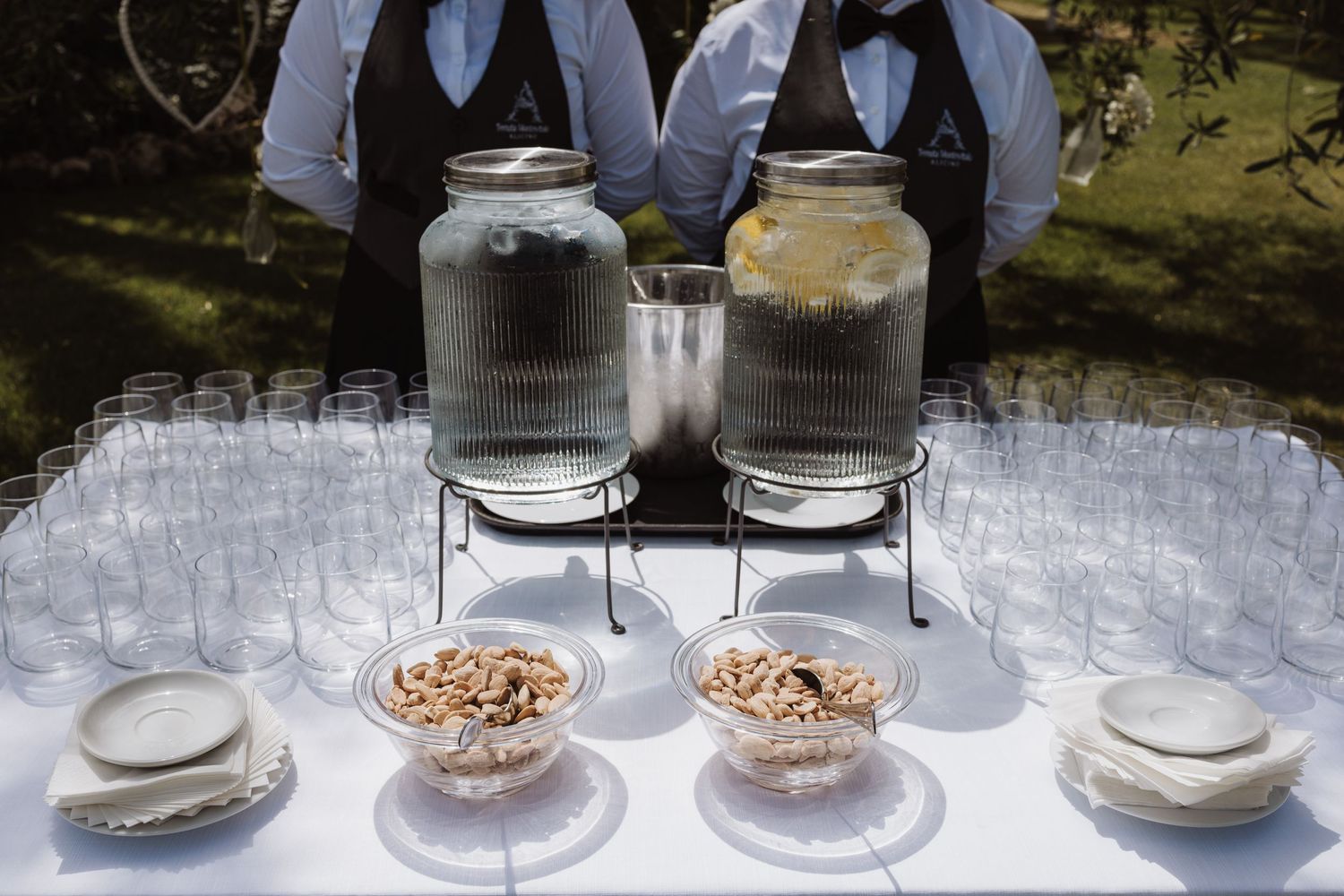 Wedding reception drink station with large glass dispensers and small bowls on white tablecloth.