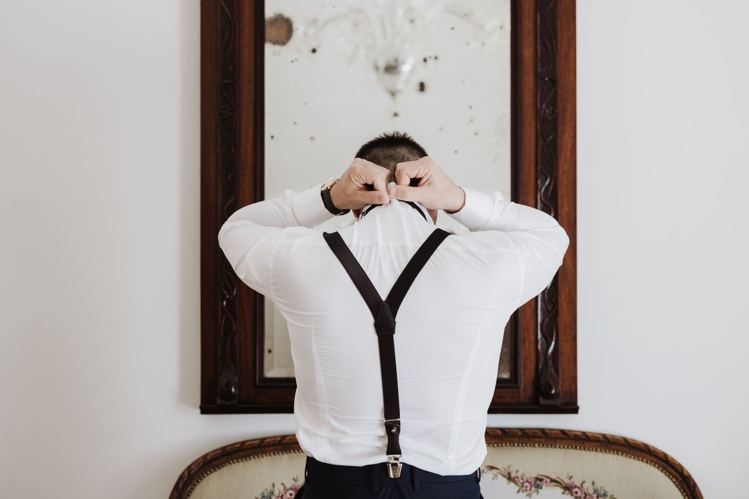 Person in white shirt and suspenders adjusts their sleeves while standing in front of a mirror.