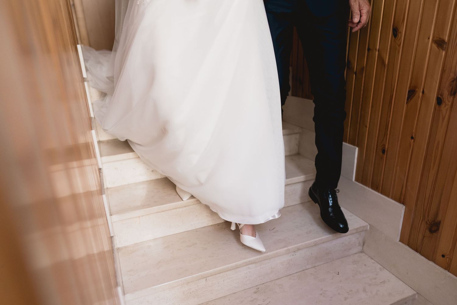 Detail shot of a flowing white wedding dress and shoes descending wooden stairs.