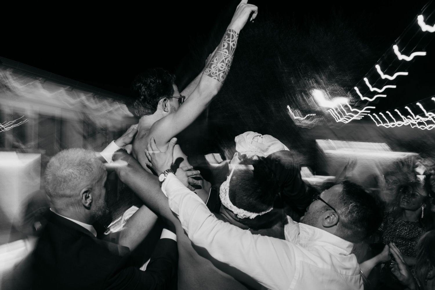 Crowd of wedding guests dancing with raised arms under twinkling string lights.