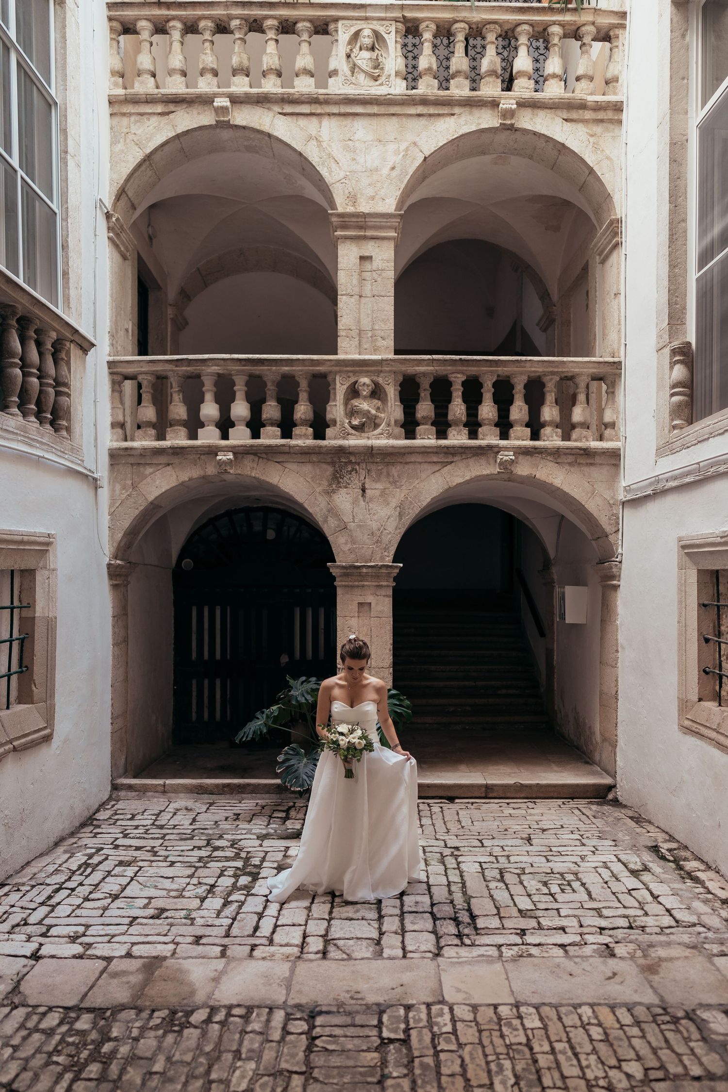 A bride in white dress standing in an ornate stone courtyard with arched architecture.