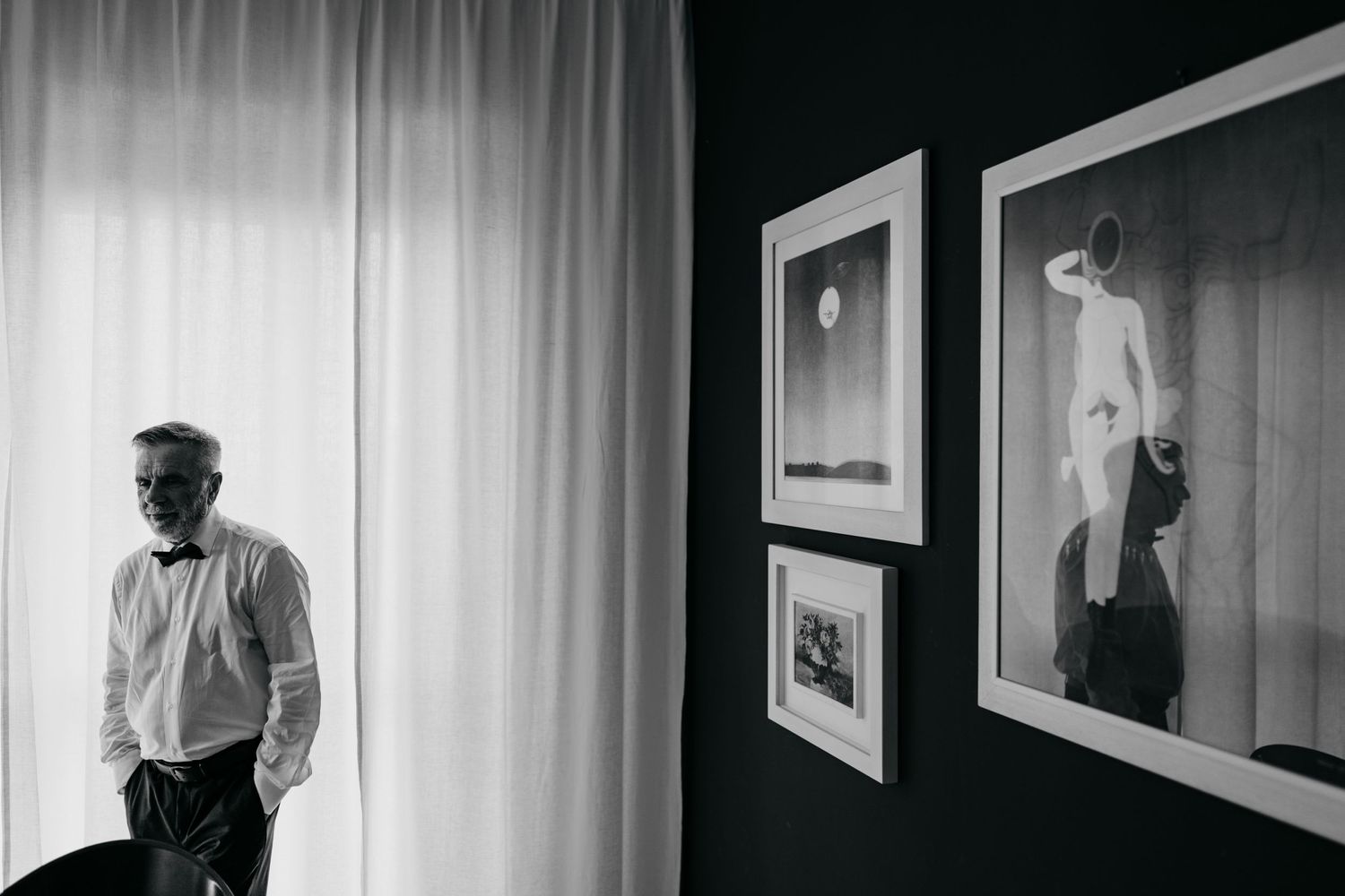 Black and white image of framed artwork hanging on a wall beside a window with curtains.
