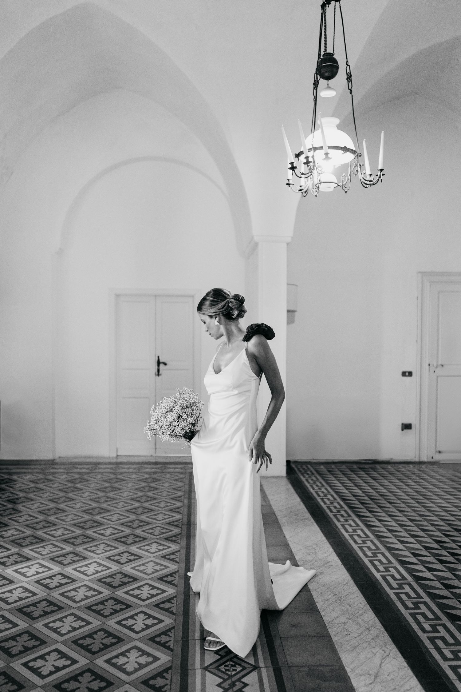 A bride in an elegant white gown stands under a vaulted ceiling with ornate tiled floors and a vintage chandelier.