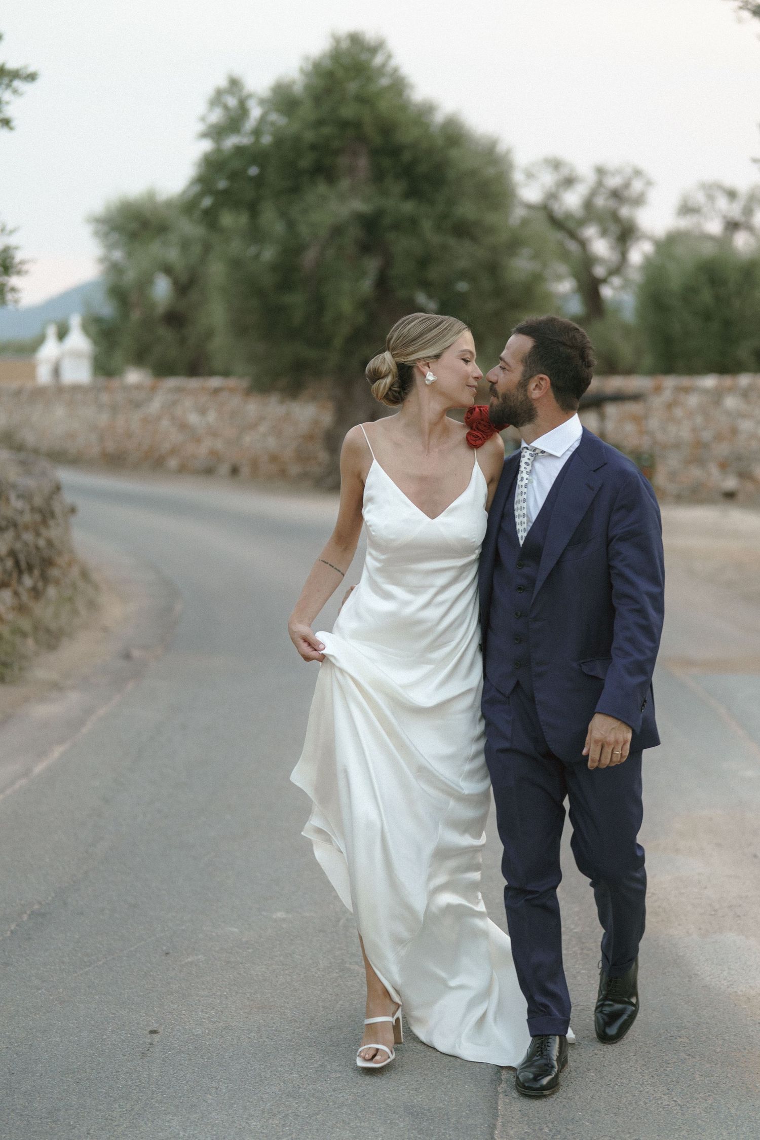 A couple in wedding attire walking together on a country road with mountains in the background.