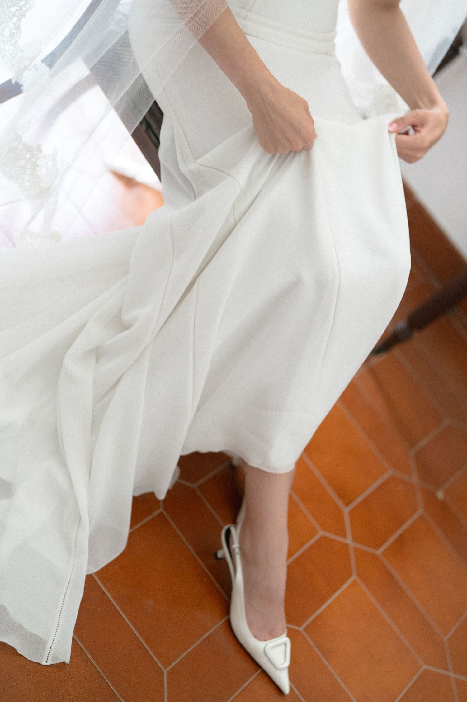 Close-up details of a white wedding dress hem and shoes on terracotta tiles.
