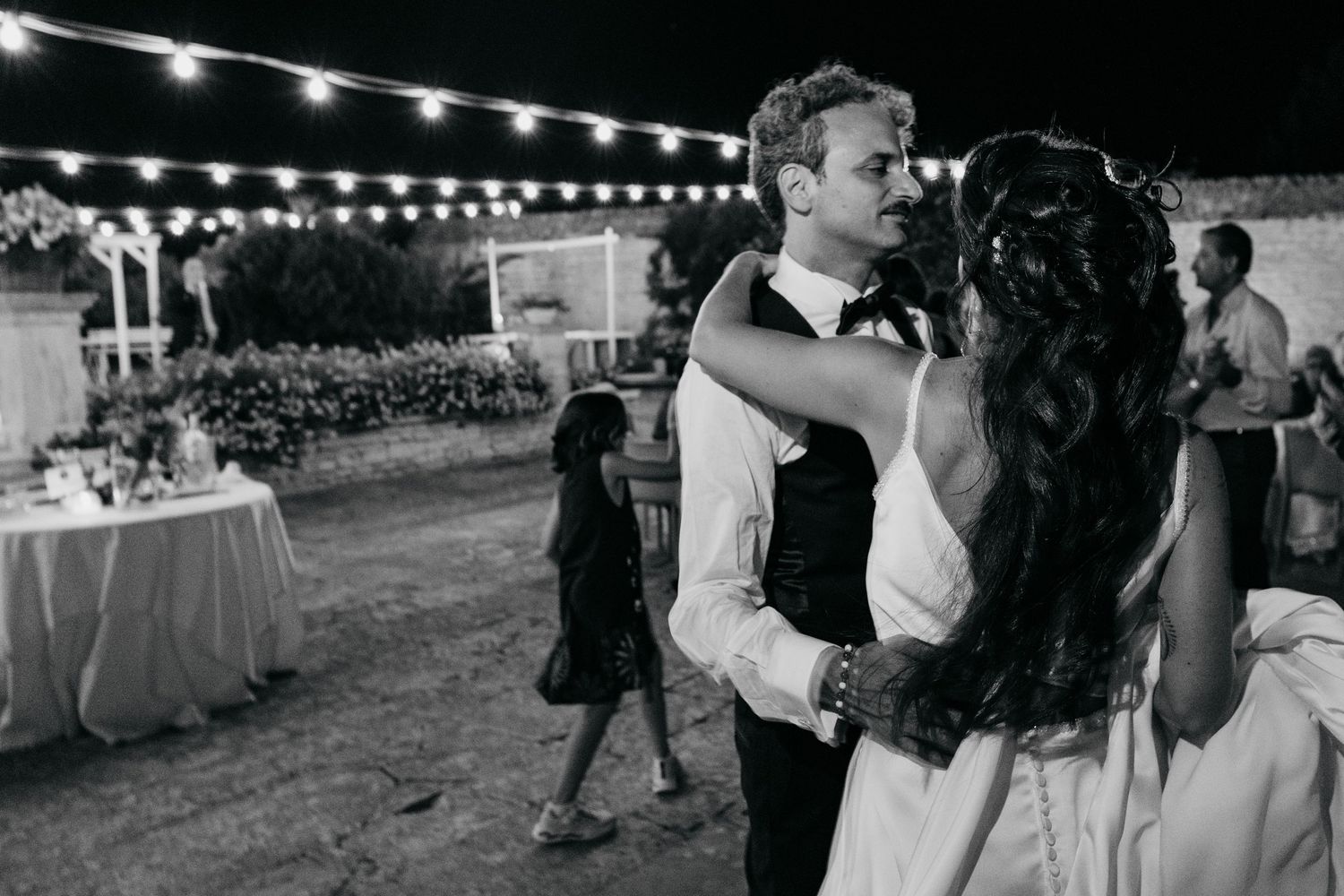 A romantic dance at an outdoor wedding reception under string lights with banquet tables in the background.