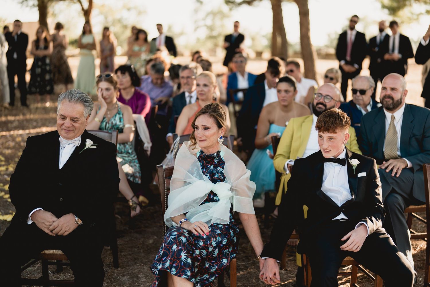 Wedding ceremony guests seated outdoors under pine trees during golden hour.