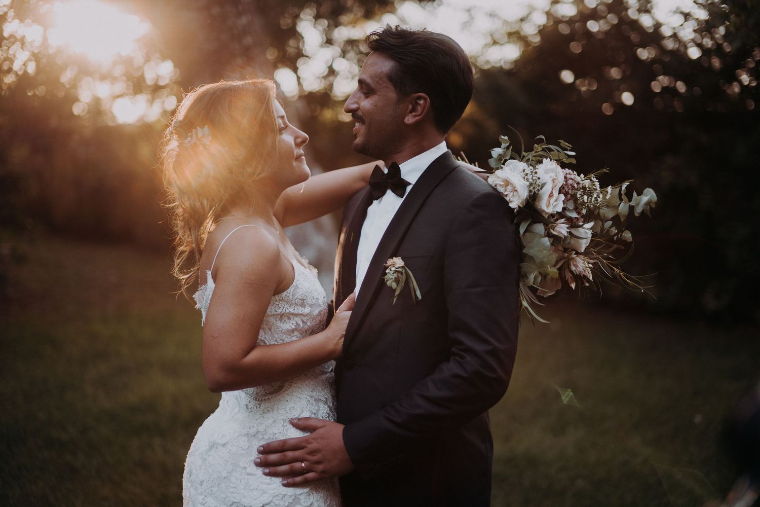 A romantic wedding couple embraces in a sunlit garden with a beautiful floral bouquet at golden hour.