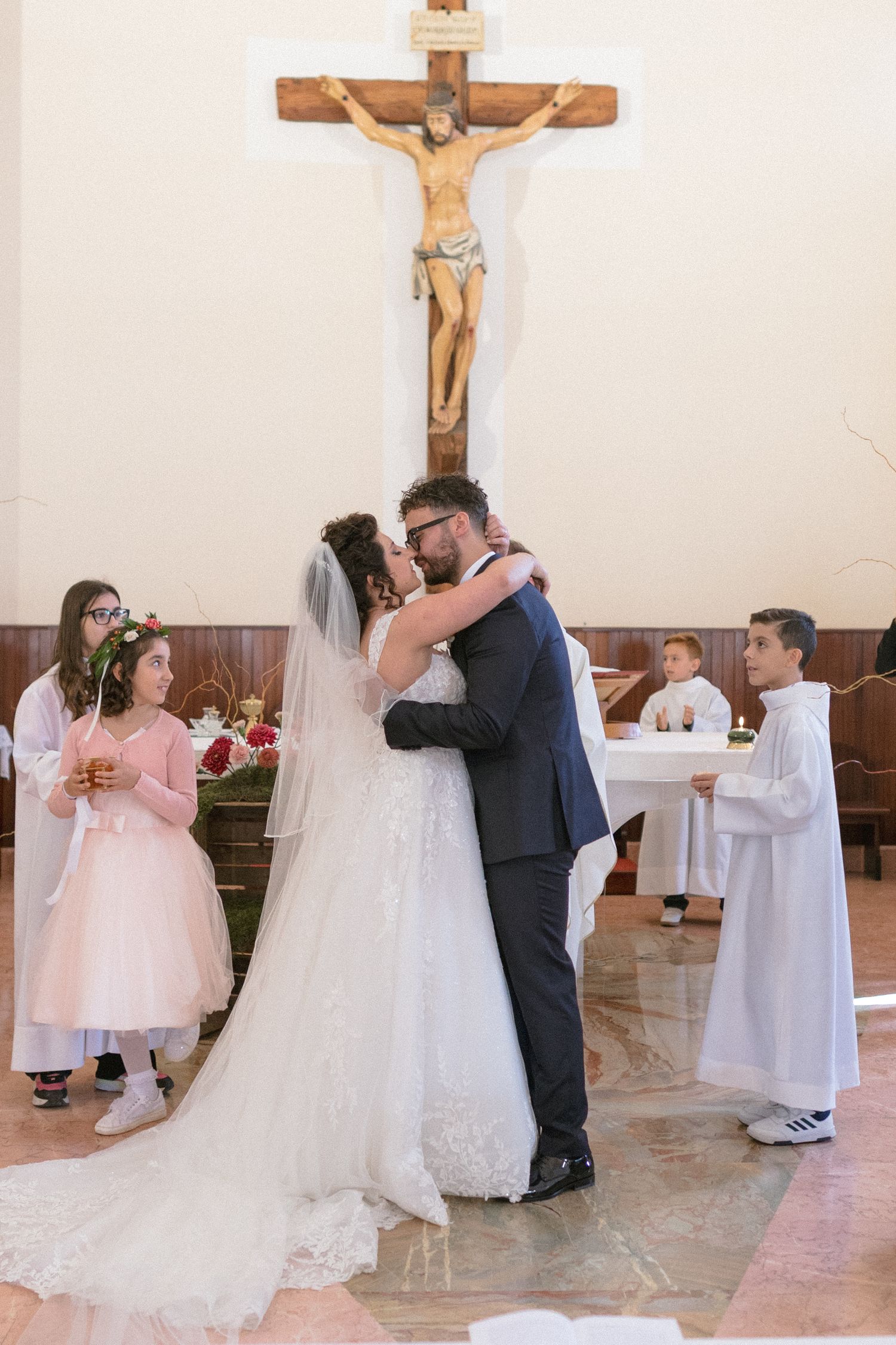 Bride and groom share first kiss at church altar with crucifix and children attendants during wedding ceremony.