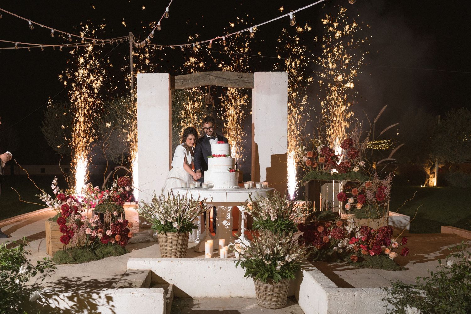 Couple cuts wedding cake under sparkling lights surrounded by colorful floral arrangements at night reception.