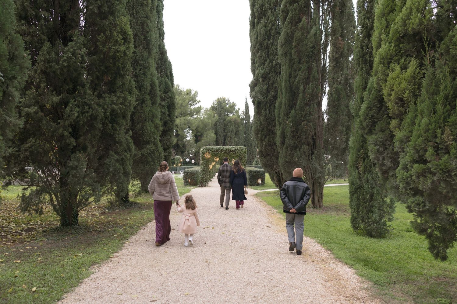 Wedding guests stroll along a gravel path lined with tall cypress trees in a scenic Italian garden.